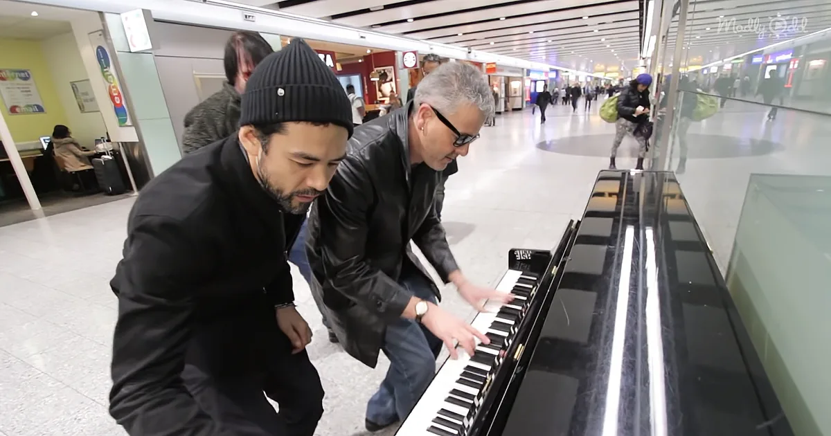 Two men, one in black beanie, lean over a piano in a busy indoor space, focusing on the keys.