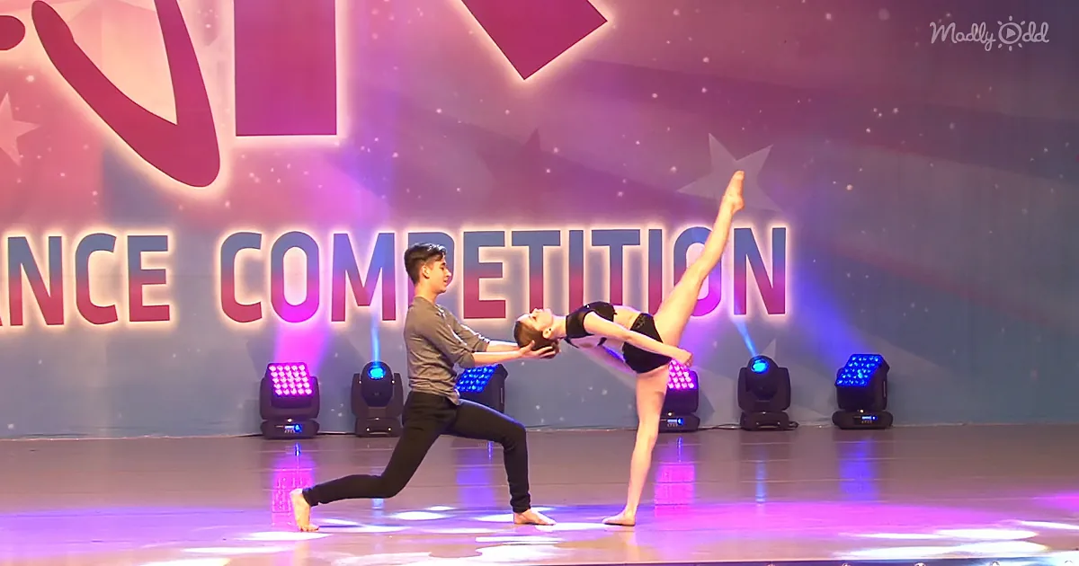 A dancer supports another in a split pose on stage at a dance competition.