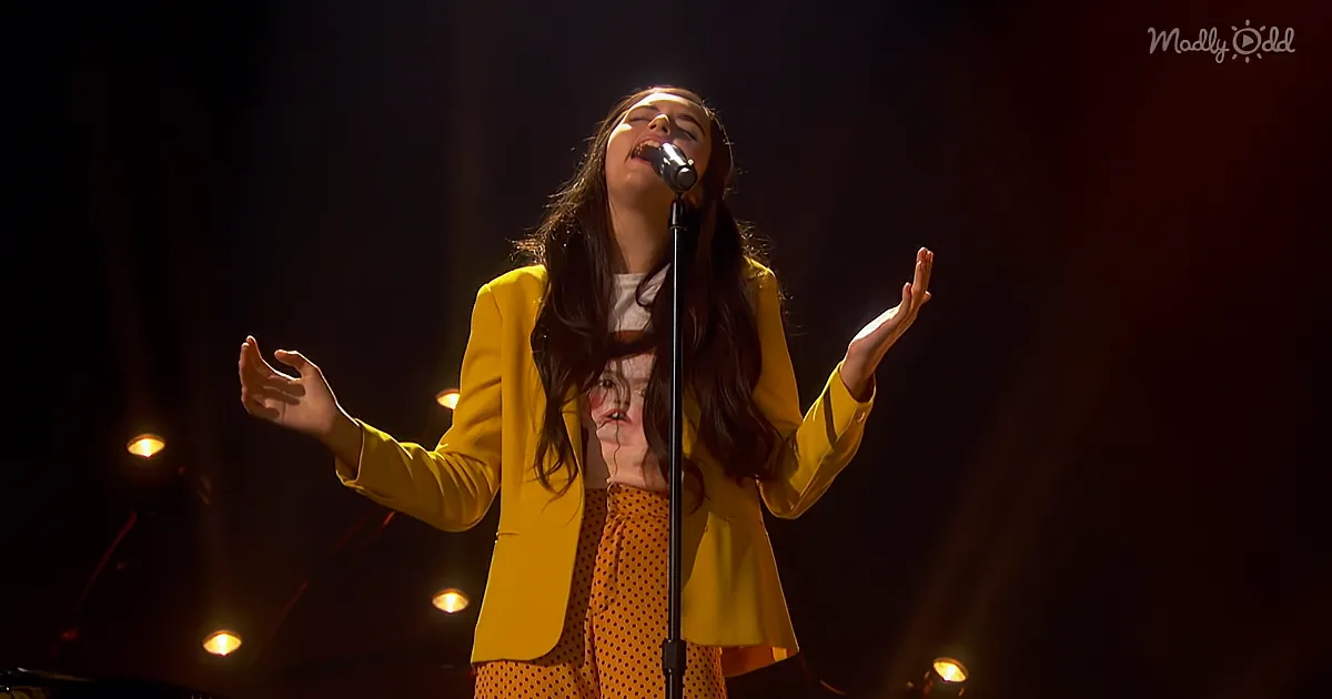 A person in a yellow outfit sings passionately on stage with arms outstretched.