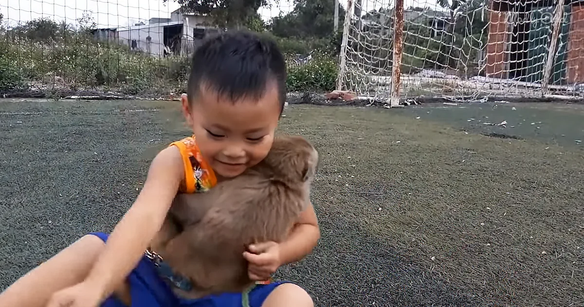 A young child in an orange shirt holds a brown stuffed animal, smiling, with a soccer goal and grassy field in the...