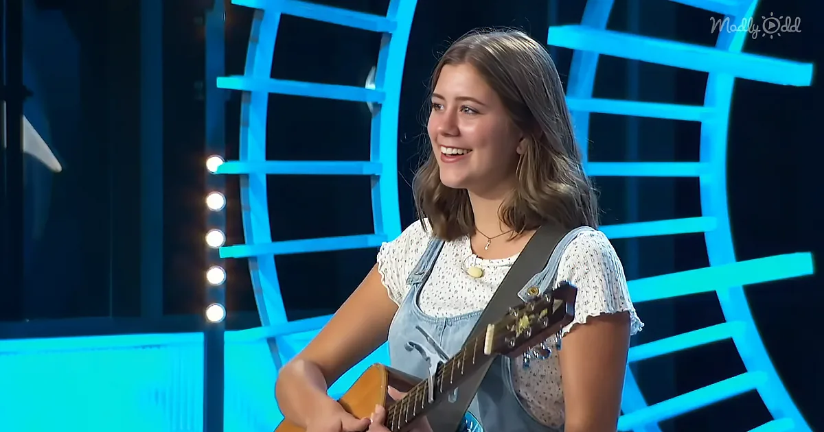 A smiling girl plays guitar on stage with blue geometric lights behind her.