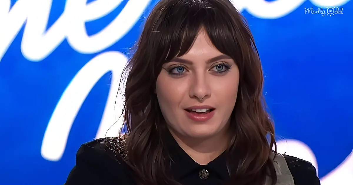 A woman with long brown hair and blue eyes, wearing a black top, is speaking in front of a blue background with white text.