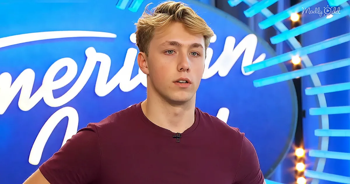 A young man in a maroon shirt stands in front of a blue "American Idol" backdrop with lights.
