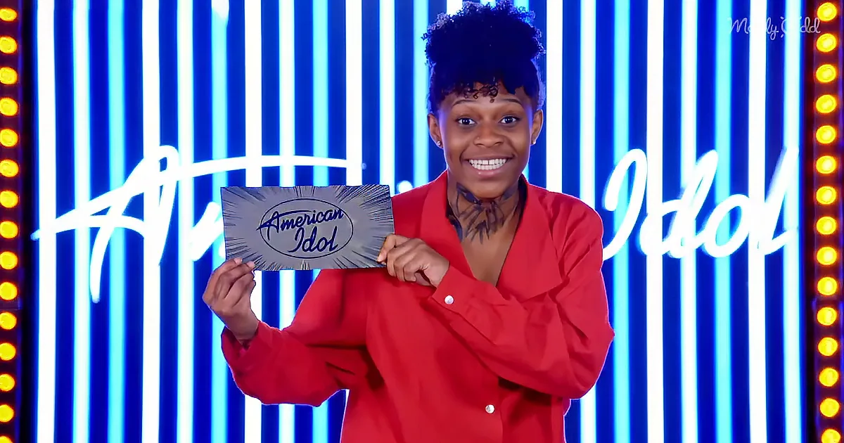 A person in a red outfit holds an "American Idol" sign against a blue and white striped background.