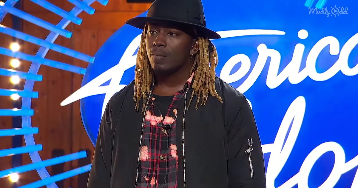 A man with dreadlocks and a hat stands in front of a blue "America's Got Talent" backdrop.