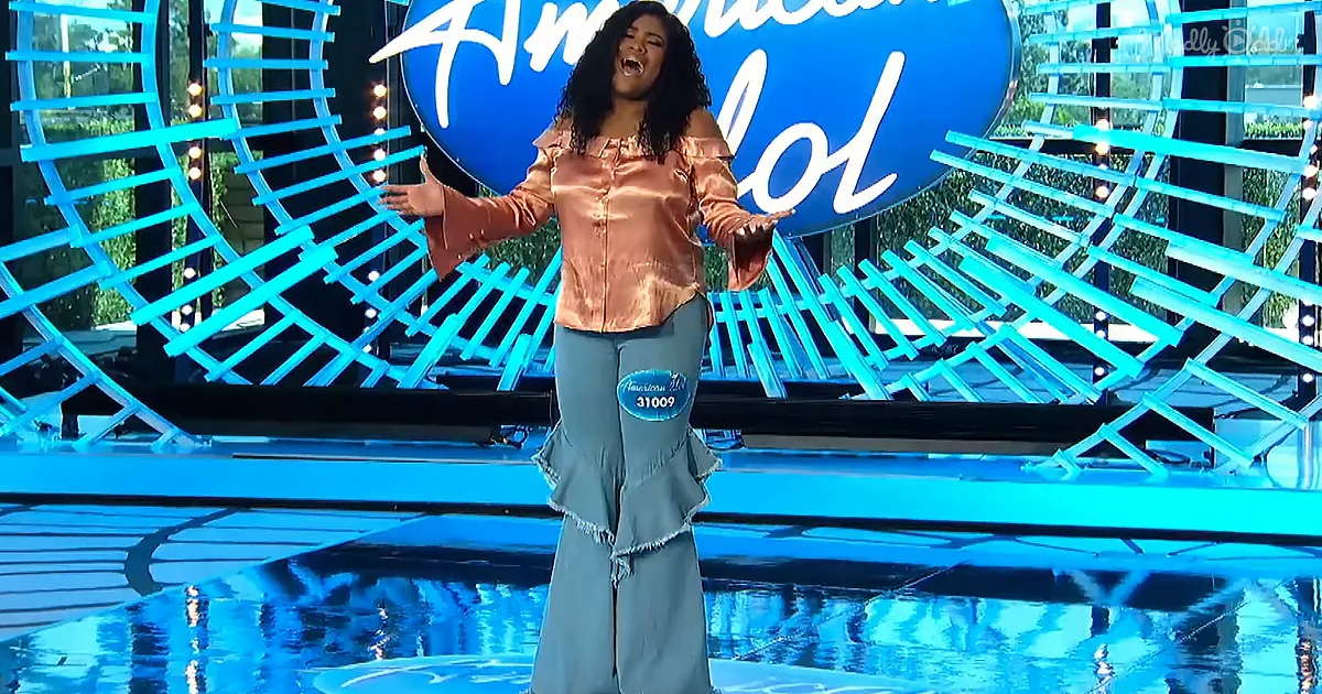 A woman in a pink top and blue flared pants stands on a stage with an "American Idol" backdrop.