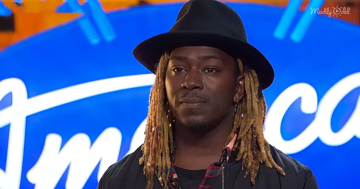 A man with long, golden dreadlocks and a black hat stands in front of a blue "America's Got Talent" backdrop.