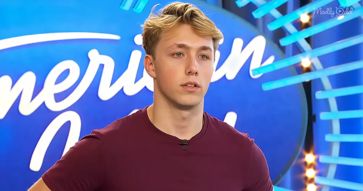 A young man in a maroon shirt stands in front of a blue "American Idol" backdrop with bright lights.