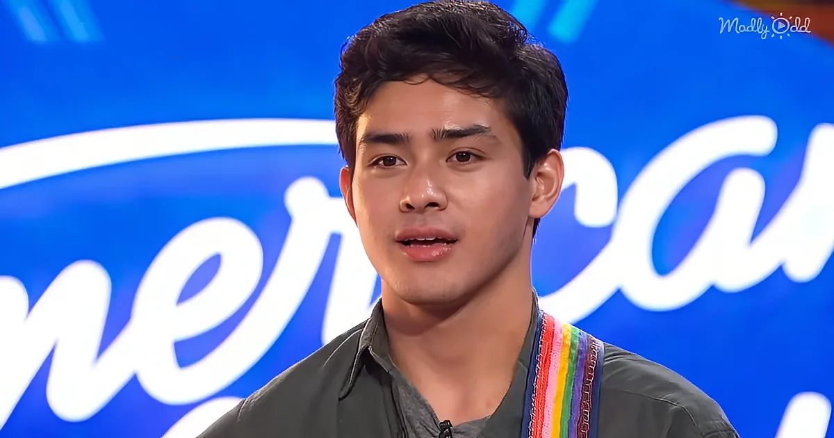 A young man with dark hair and a gray shirt stands in front of a blue background with "American" partially visible.