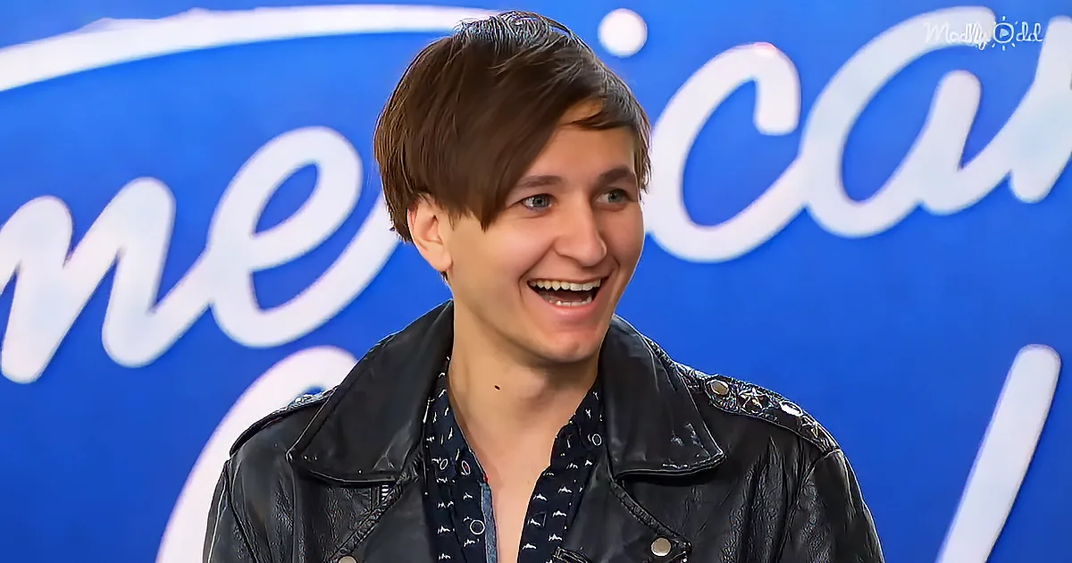 A person in a black leather jacket smiles in front of a blue "American Idol" backdrop.