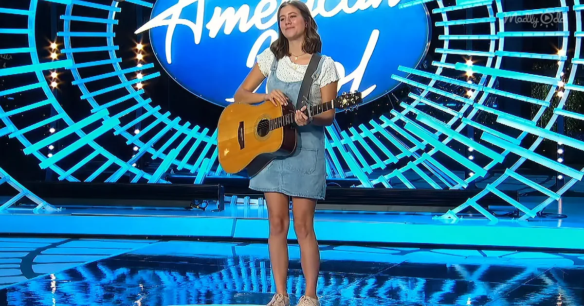 A young woman in a denim dress holds a guitar on a stage with a blue "American Idol" backdrop.