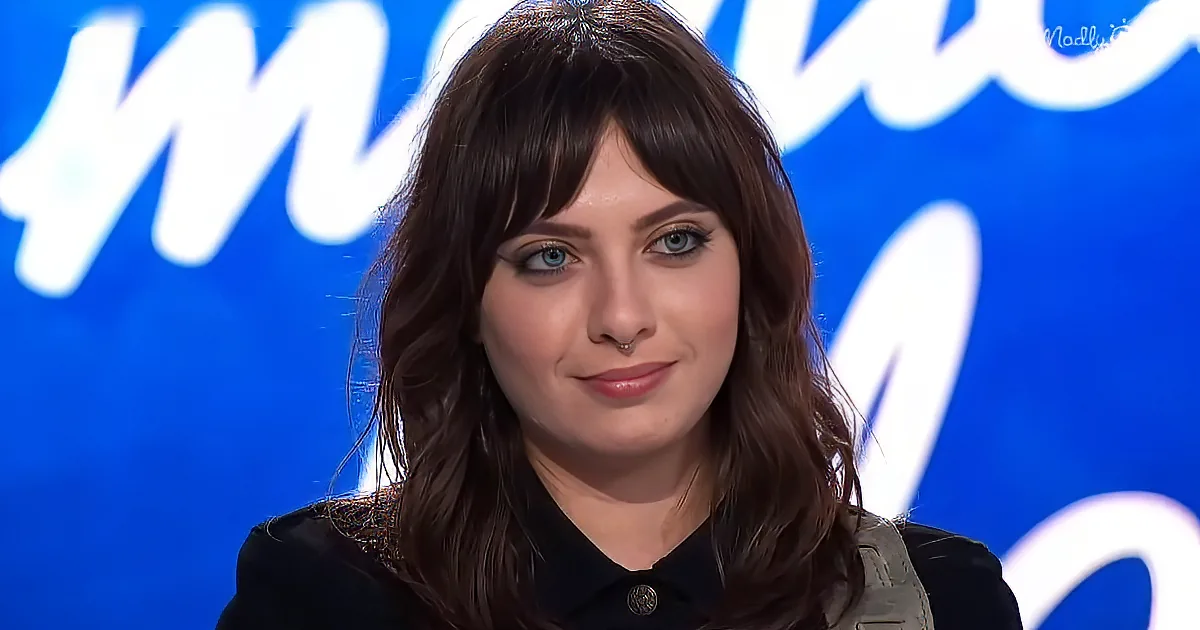 A person with long brown hair and a nose ring is wearing a black shirt, sitting in front of a blue background with white...