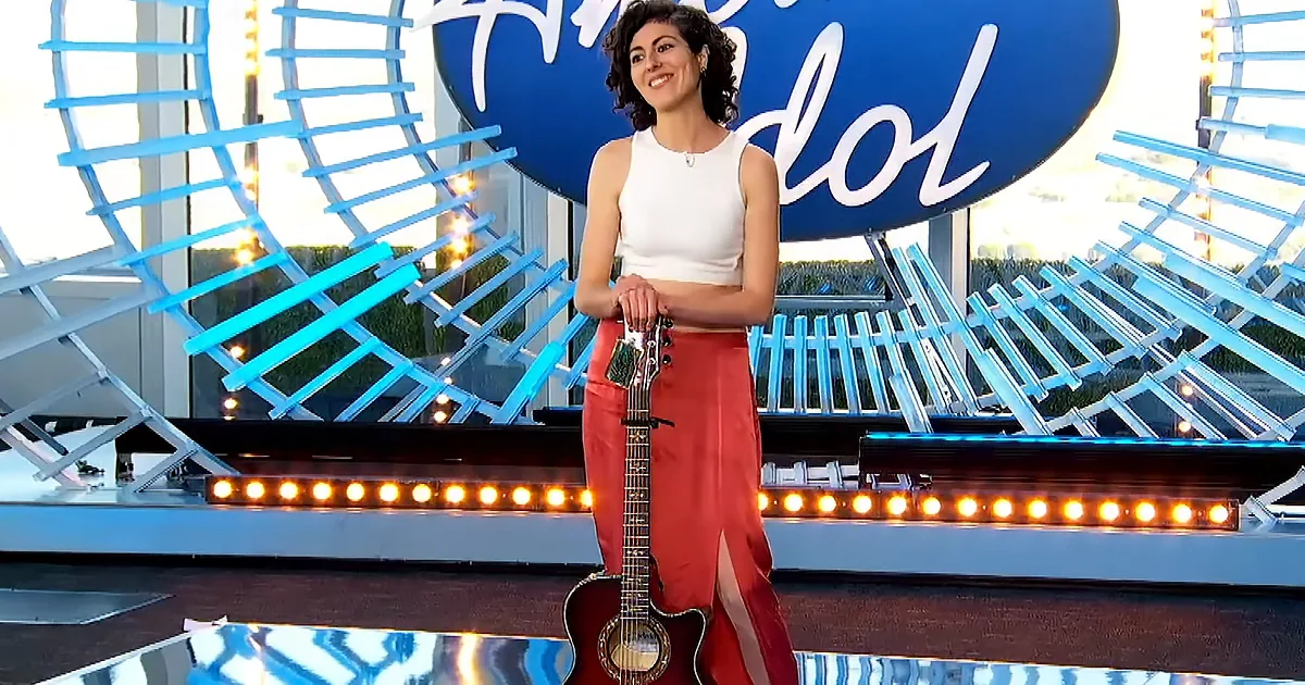 A woman in a white top and red skirt stands holding a guitar on a stage with "Idol" in the background.