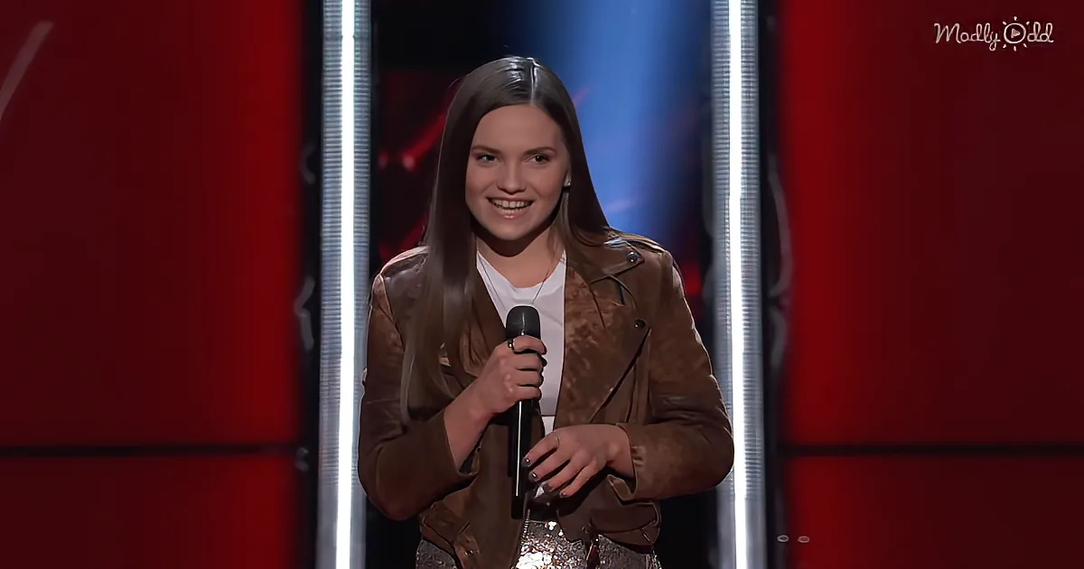 A person in a brown jacket holds a microphone on stage with red and white lights.