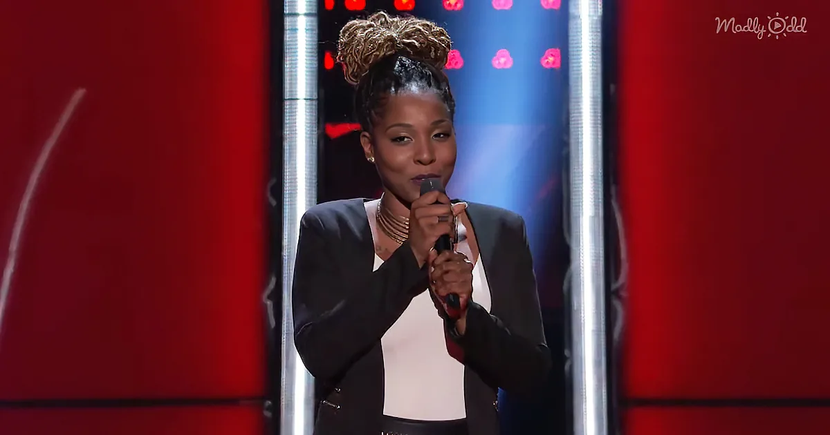 A woman with curly hair in a bun, wearing a black blazer and white top, holds a microphone on stage with red and blue...