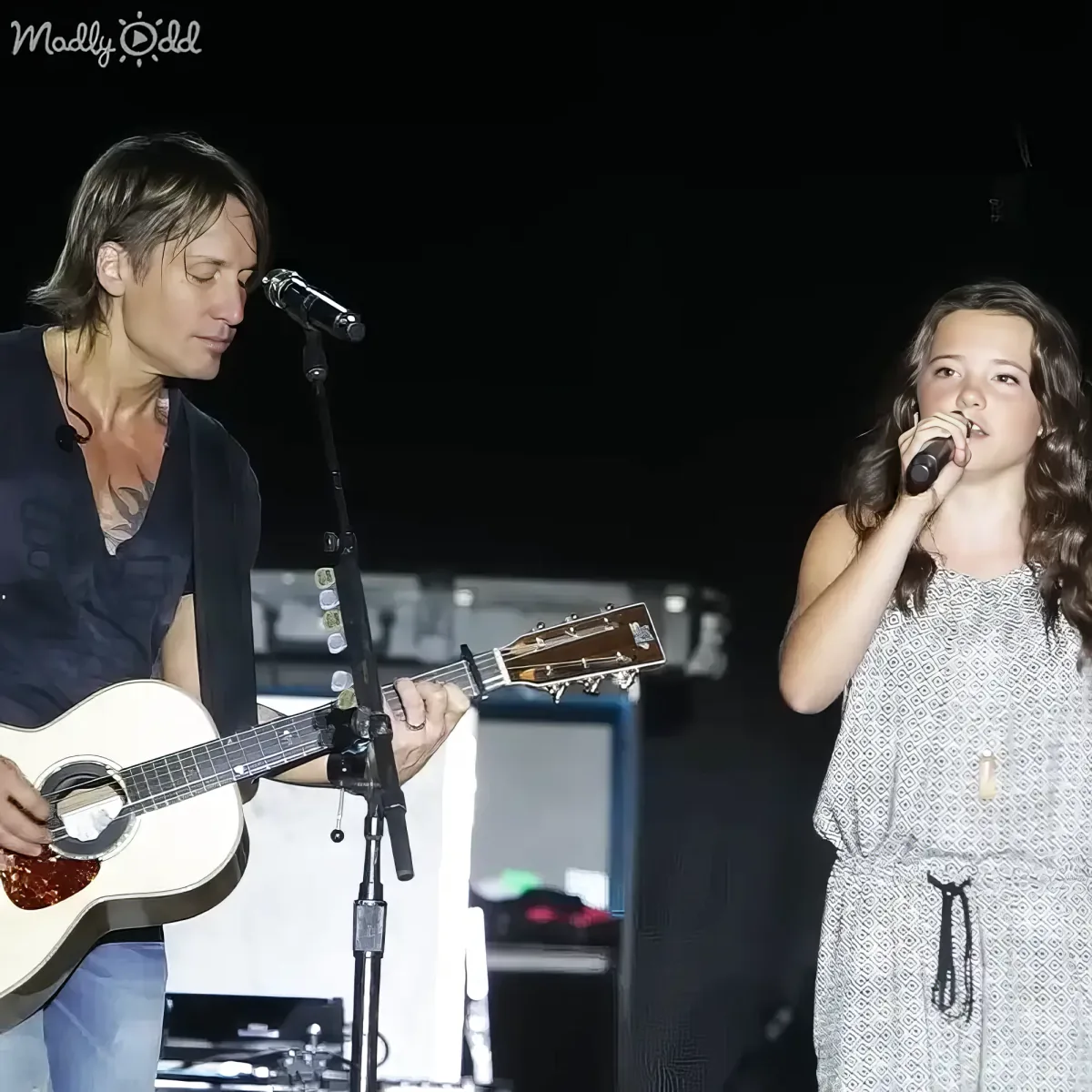 A man plays guitar while a young girl sings into a microphone on stage.