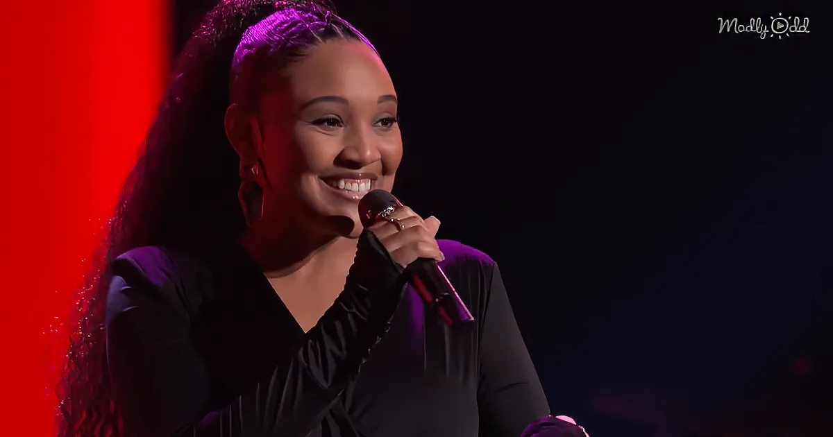 A woman with long hair, wearing a black outfit, smiles while holding a microphone on stage.