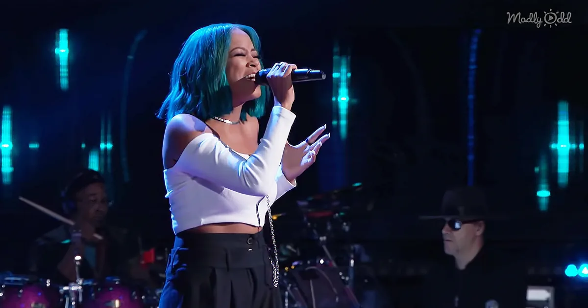 A performer with blue hair sings into a microphone, wearing a white top and black pants, with a band in the background.