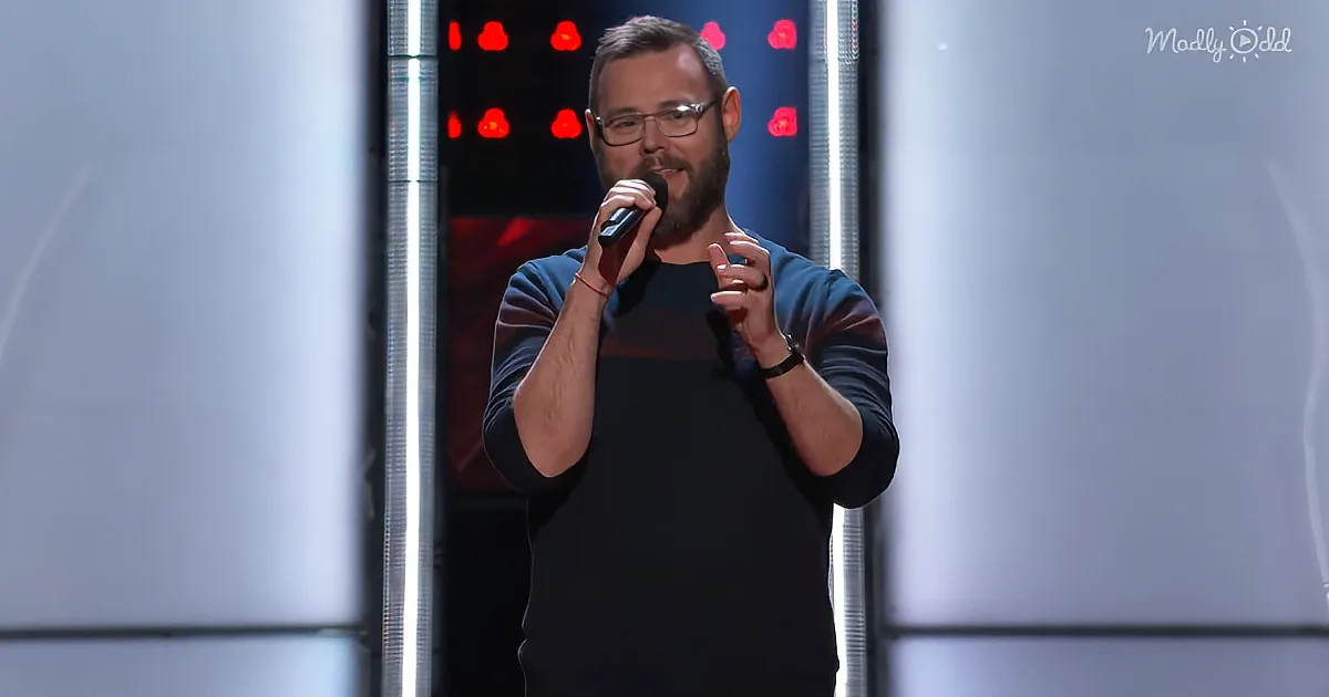 A man with glasses and a beard holds a microphone, gesturing with his other hand on stage.
