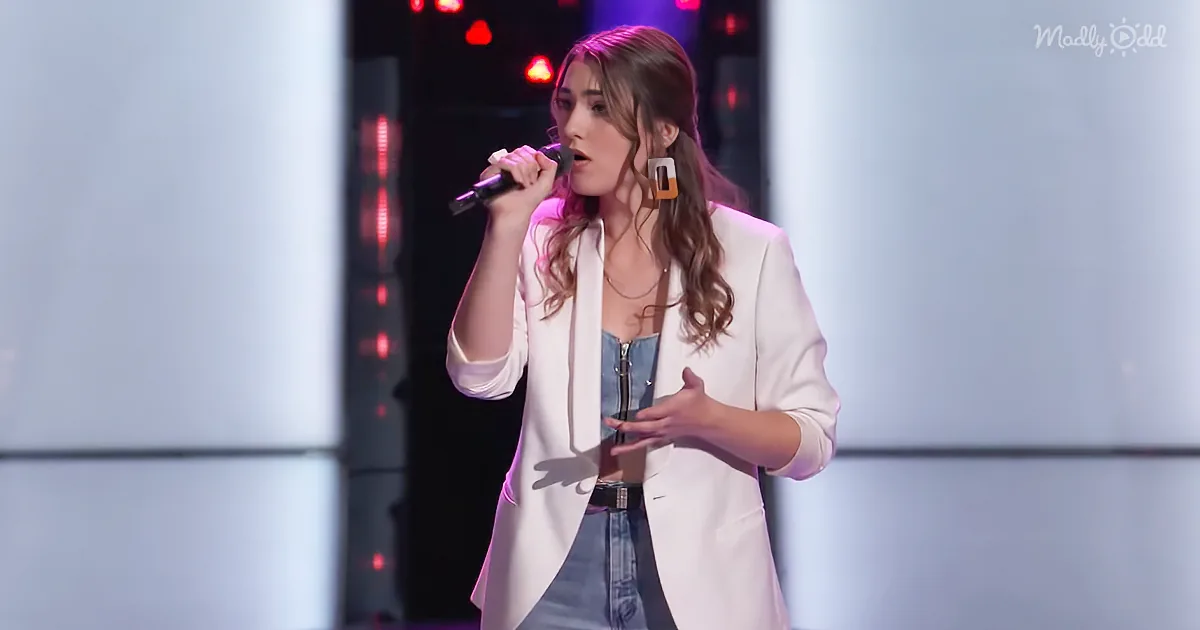A woman in a white blazer and jeans sings into a microphone on stage with red lights in the background.