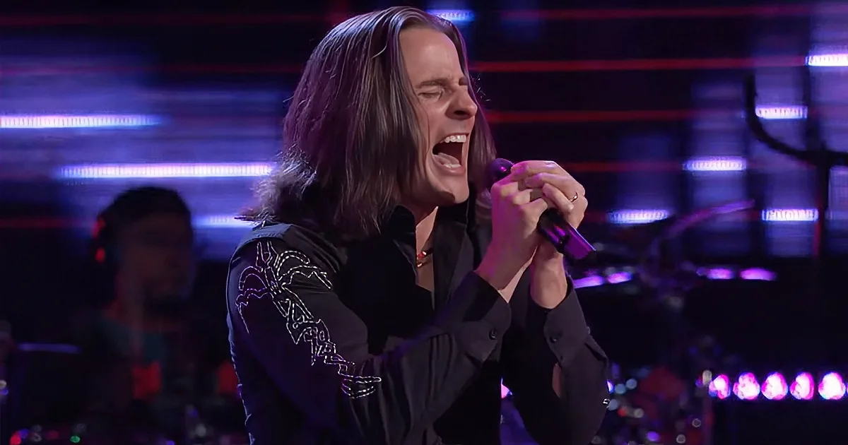 A man with long hair sings passionately into a microphone on stage.