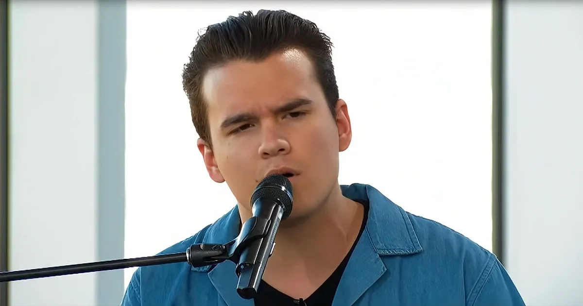 A man in a blue shirt sings into a microphone, his mouth open, against a bright background.