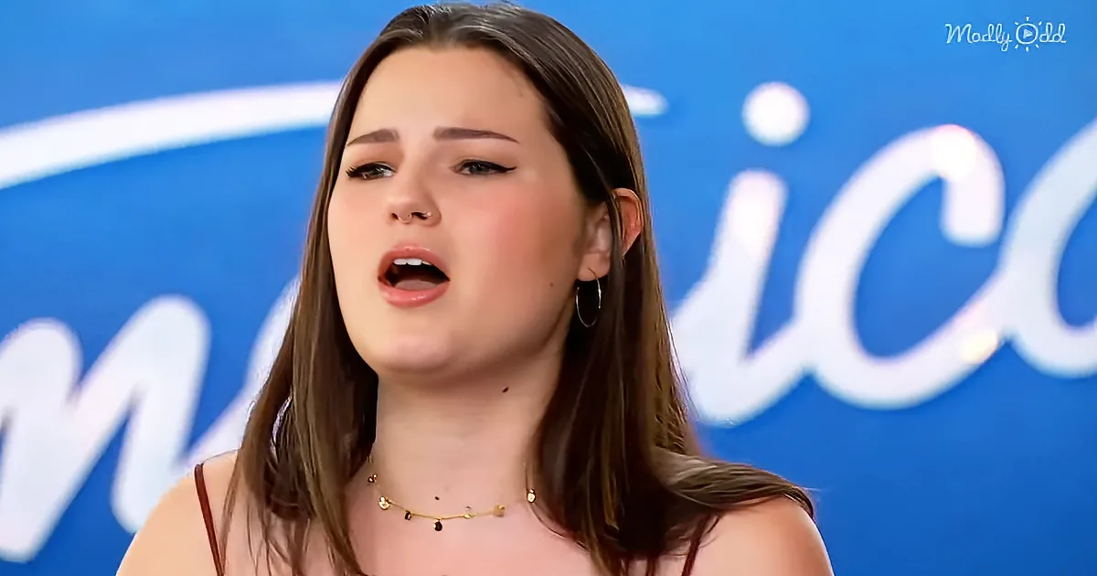 A young woman with long brown hair and a nose ring is singing on stage, wearing a gold necklace and hoop earrings.