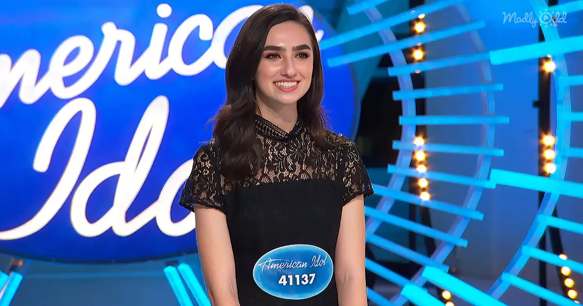 A smiling woman in a black lace dress stands on an "American Idol" stage, wearing a contestant badge.