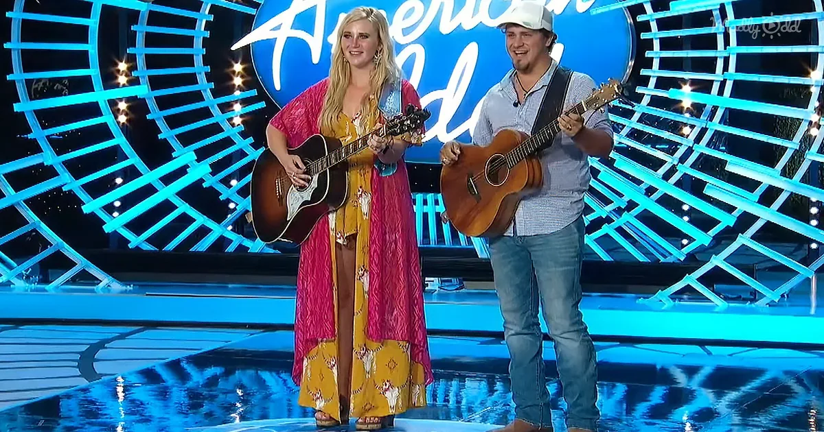 A man and woman stand on stage, each holding an acoustic guitar, in front of a blue "American Idol" backdrop.