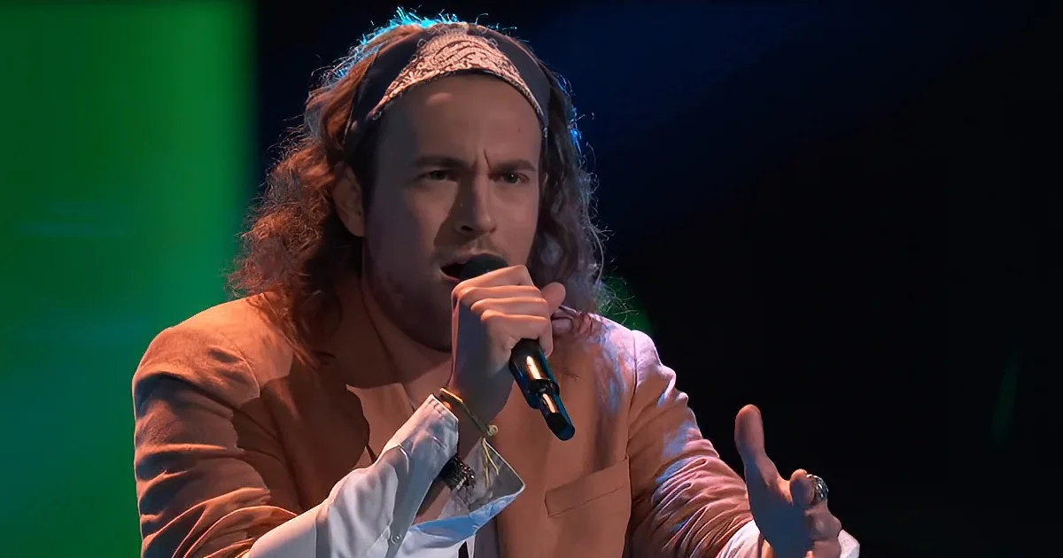 A person with long hair and a headband sings into a microphone on stage, wearing a light-colored jacket.