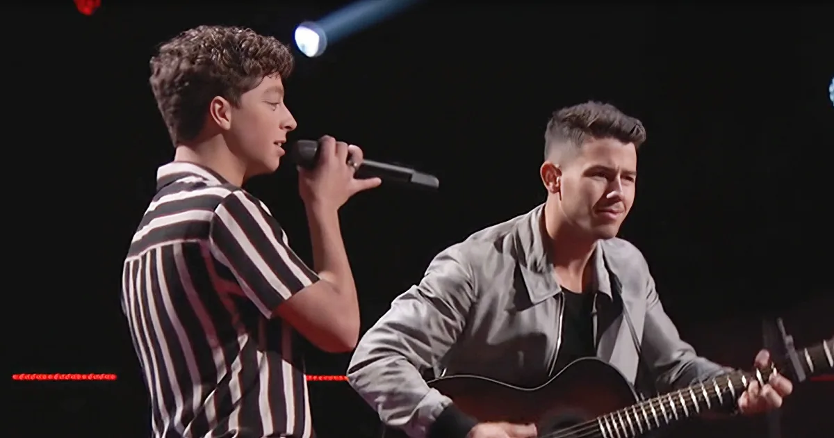 A young man sings into a microphone while another plays guitar on stage.