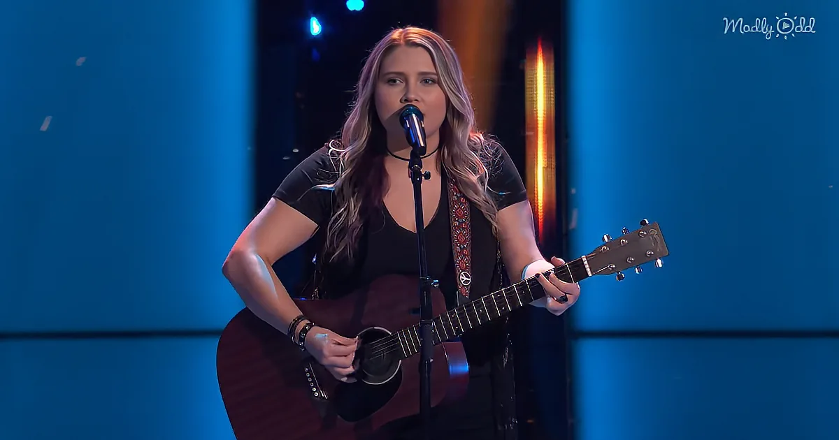 A woman with long hair plays an acoustic guitar and sings into a microphone on stage.