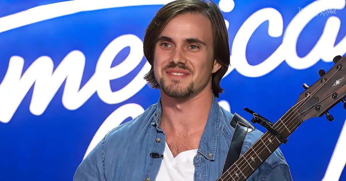 A man with long hair and a beard smiles while holding a guitar, standing in front of a blue "America's Got Talent" backdrop.