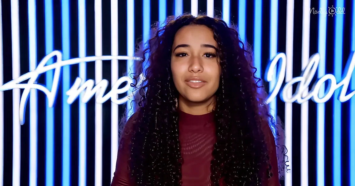 A person with long, curly hair stands in front of a backdrop with "American Idol" written in white cursive.