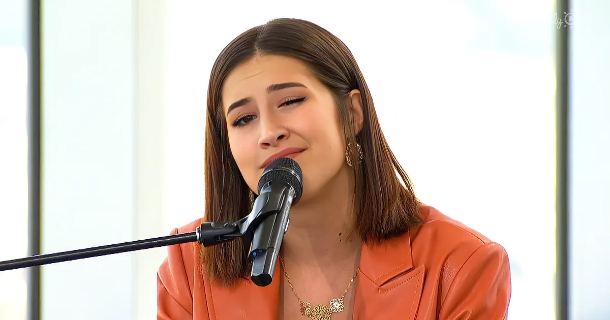 A woman in an orange jacket sings into a microphone, her eyes closed, with a gold necklace and earrings.
