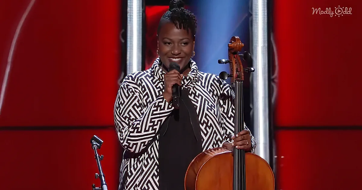 A woman in a black-and-white patterned coat holds a cello and a microphone on stage.