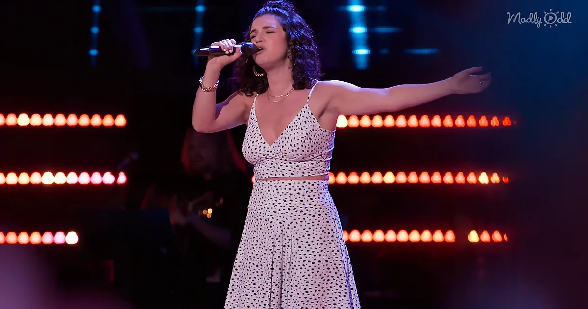A woman in a polka-dot dress sings passionately on stage with red lights in the background.