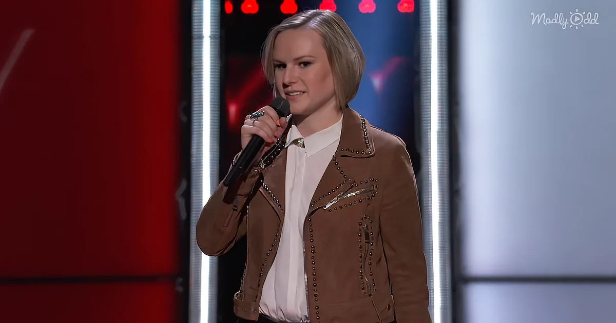 A person in a brown studded jacket holds a microphone on stage with red and white lights behind.