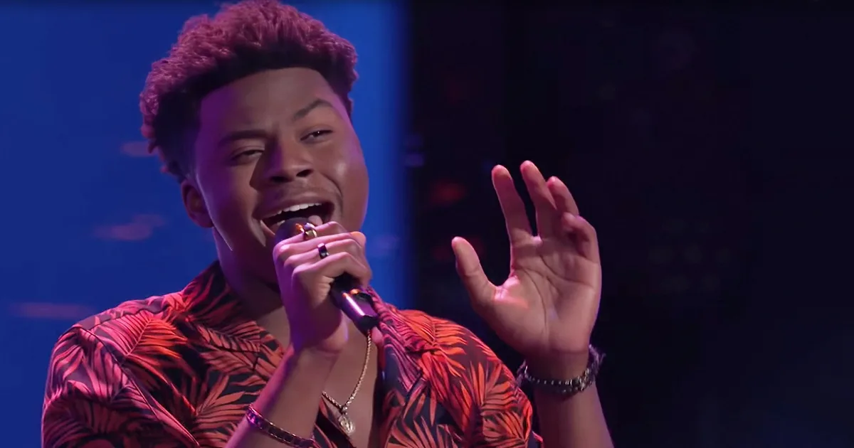 A person in a floral shirt sings into a microphone, gesturing with their hands.