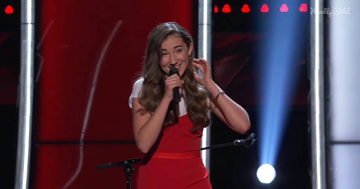 A woman in a red dress holds a microphone on stage, smiling with her hand near her ear.