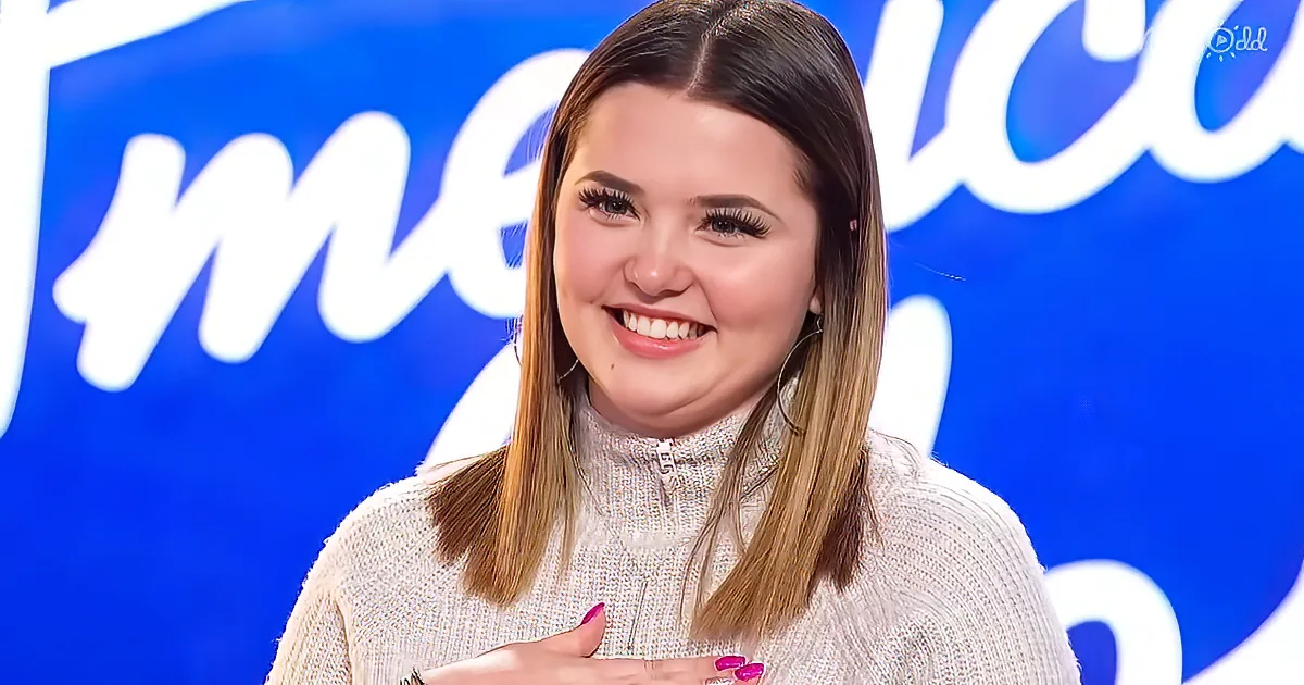 A smiling woman with long hair, wearing a white sweater, and hoop earrings, stands in front of a blue background with...