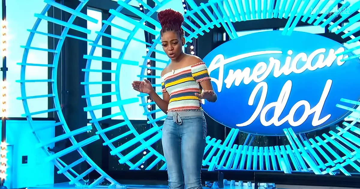 A woman stands on stage in front of an "American Idol" sign, gesturing with her hands.