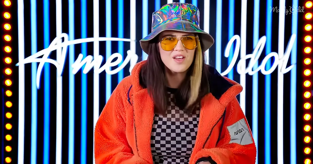 A person wearing a reflective hat, orange jacket, and checkered shirt stands in front of a neon-lit "American Idol" backdrop.