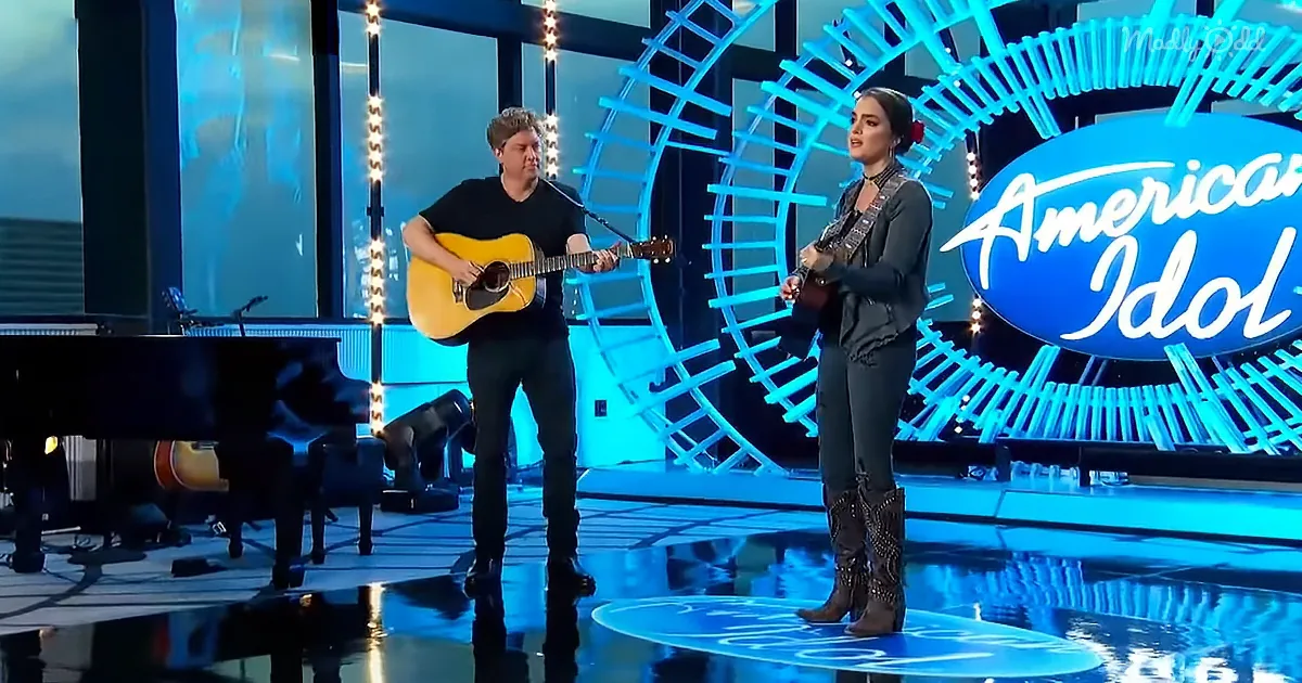 A man and woman perform on stage with guitars, "American Idol" logo in the background.