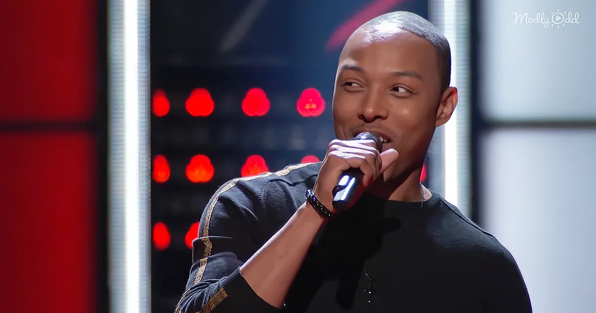 A man in a black shirt sings into a microphone on a stage with red lights behind him.
