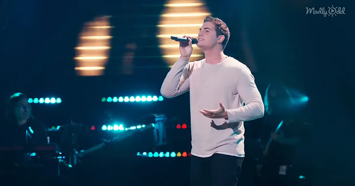 A man in a white shirt sings into a microphone on stage with colorful lights behind him.