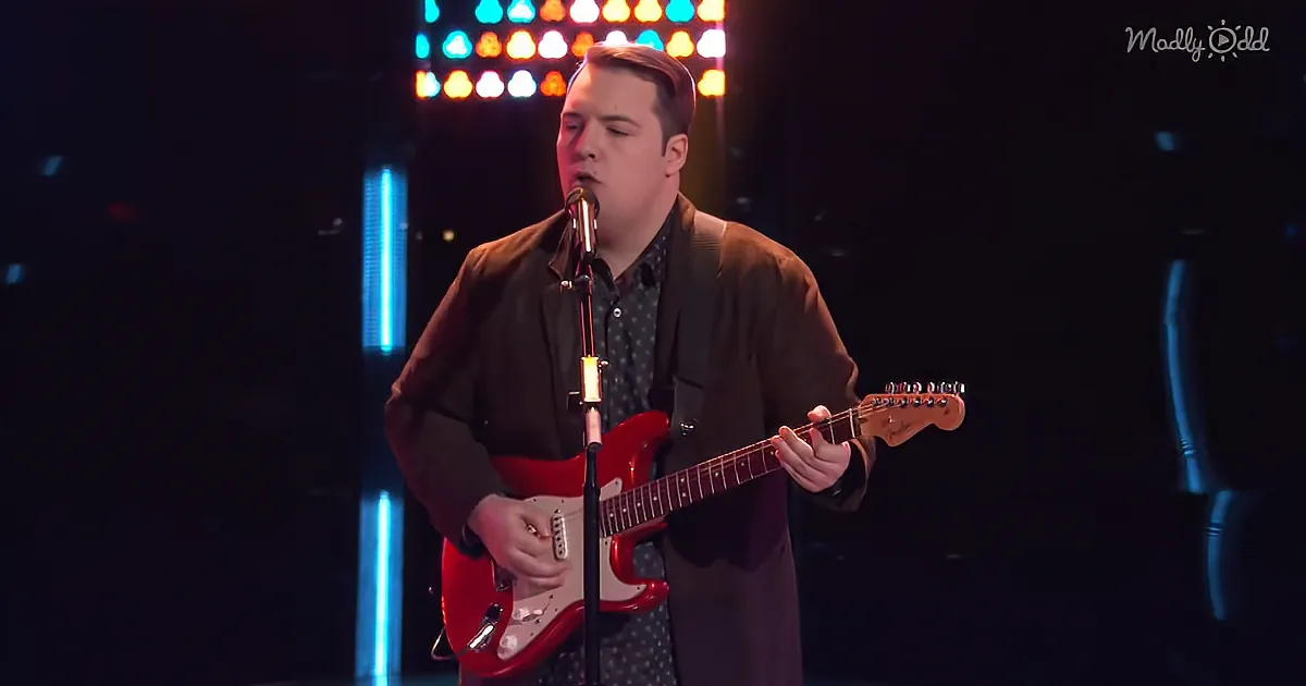 A man in a suit plays a red electric guitar on stage with a microphone.