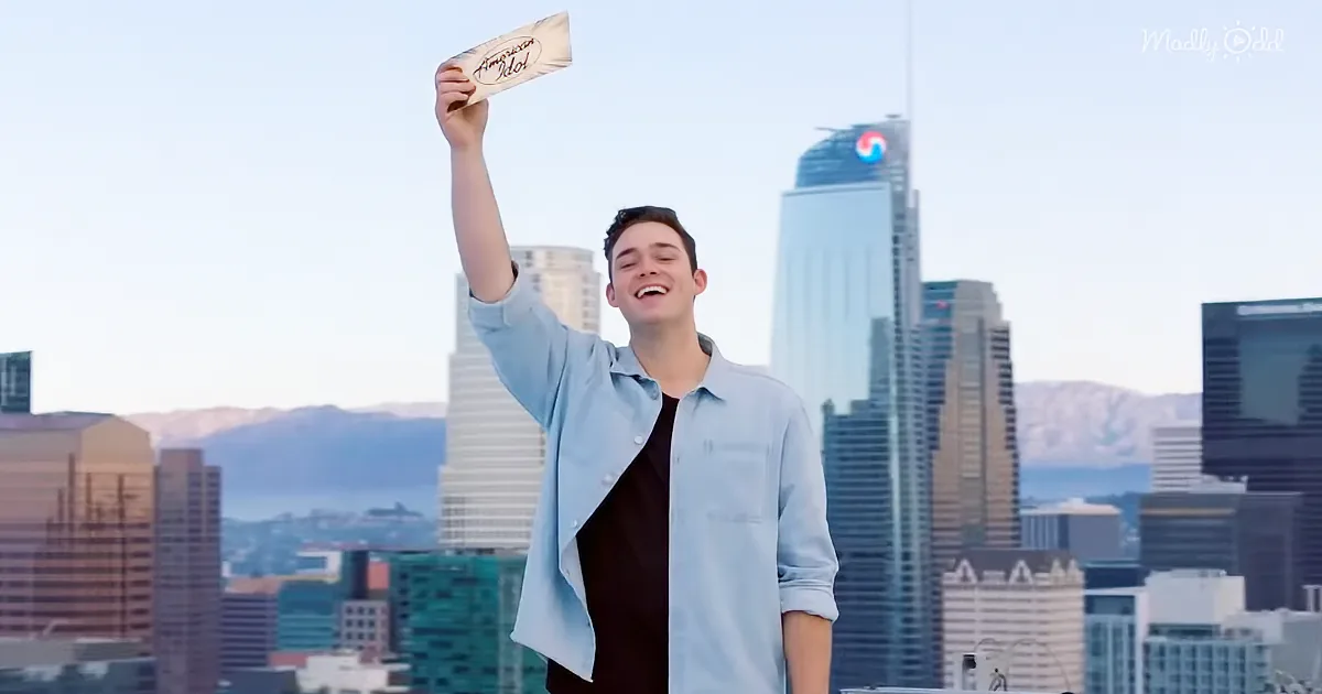 A man in a light blue shirt holds up a sign with "American Idol" written on it, standing in front of a city skyline.