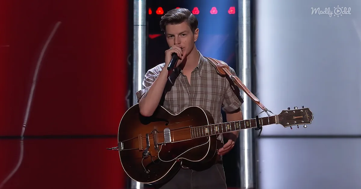 A young man in a plaid shirt holds a microphone and a guitar on stage.