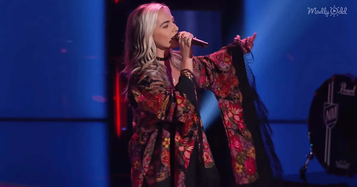 A woman in a floral robe sings into a microphone on stage, illuminated by blue lights.